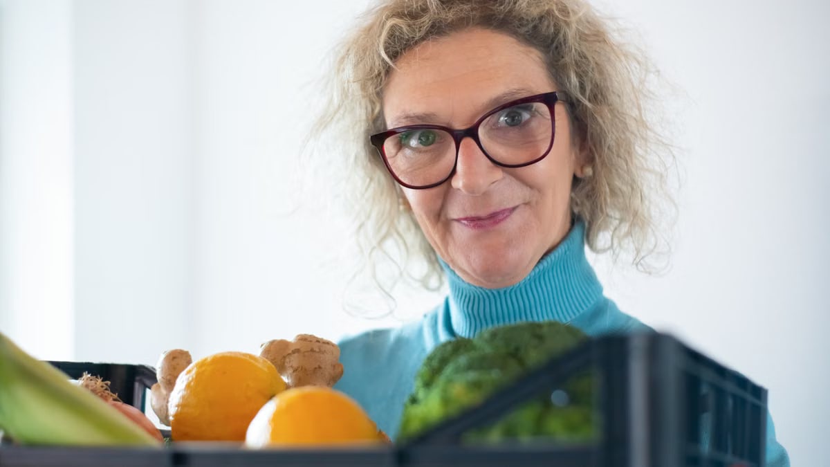 Elderly woman smiling and holding fresh fruit and vegetables indoors.